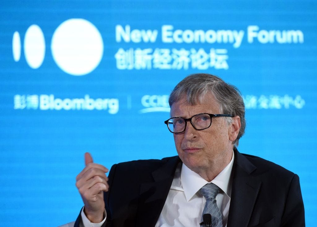 Billionaire Bill Gates sits on stage in front of a blue background wearing a black suit jacket and grey tie.